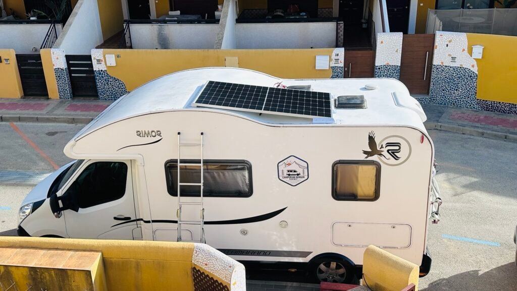 White motorhome with a large rooftop solar panel, ladder on the side, and a bird logo, parked in front of a yellow building with mosaic accents.