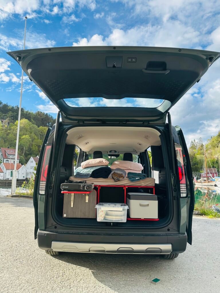 Open SUV trunk filled with blankets, pillows, and storage bins for a road trip or camping setup, outdoors near a marina.