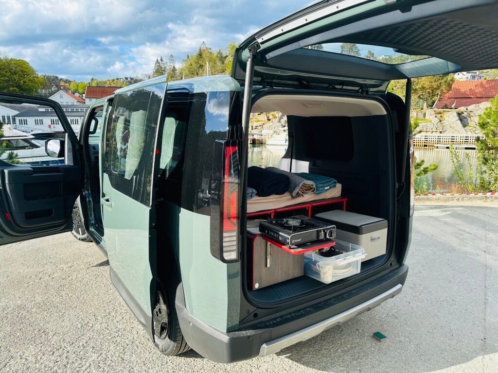 Open-back camper van with organized storage drawers, blankets, and gear visible inside by the waterfront.