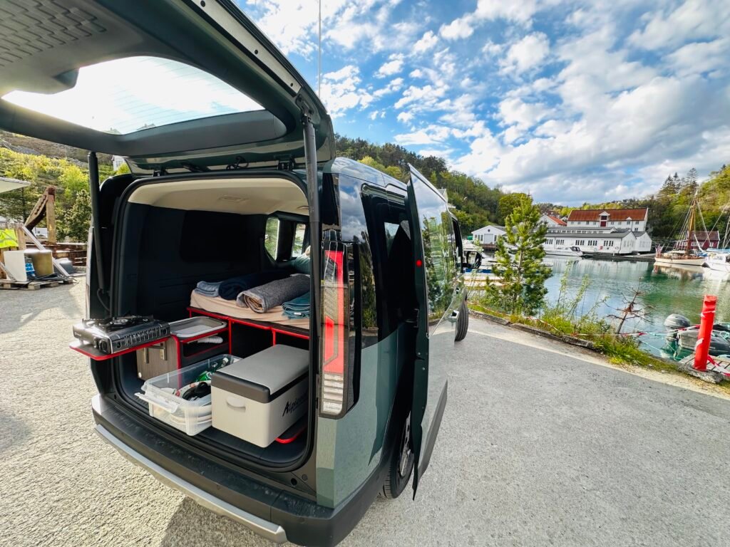 Open van showing a built-out camper setup with a pull-out kitchen, drawers, and storage in a marina town backdrop with boats and white buildings.