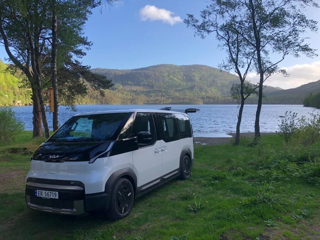 White van parked on grassy lakeshore, with calm blue water, forested mountains in the background and trees framing the scene