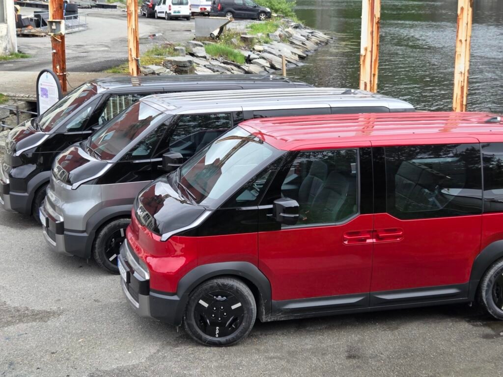 Row of dark gray, red, and black vans parked along a waterfront with a rocky embankment and rusted metal posts in the background.