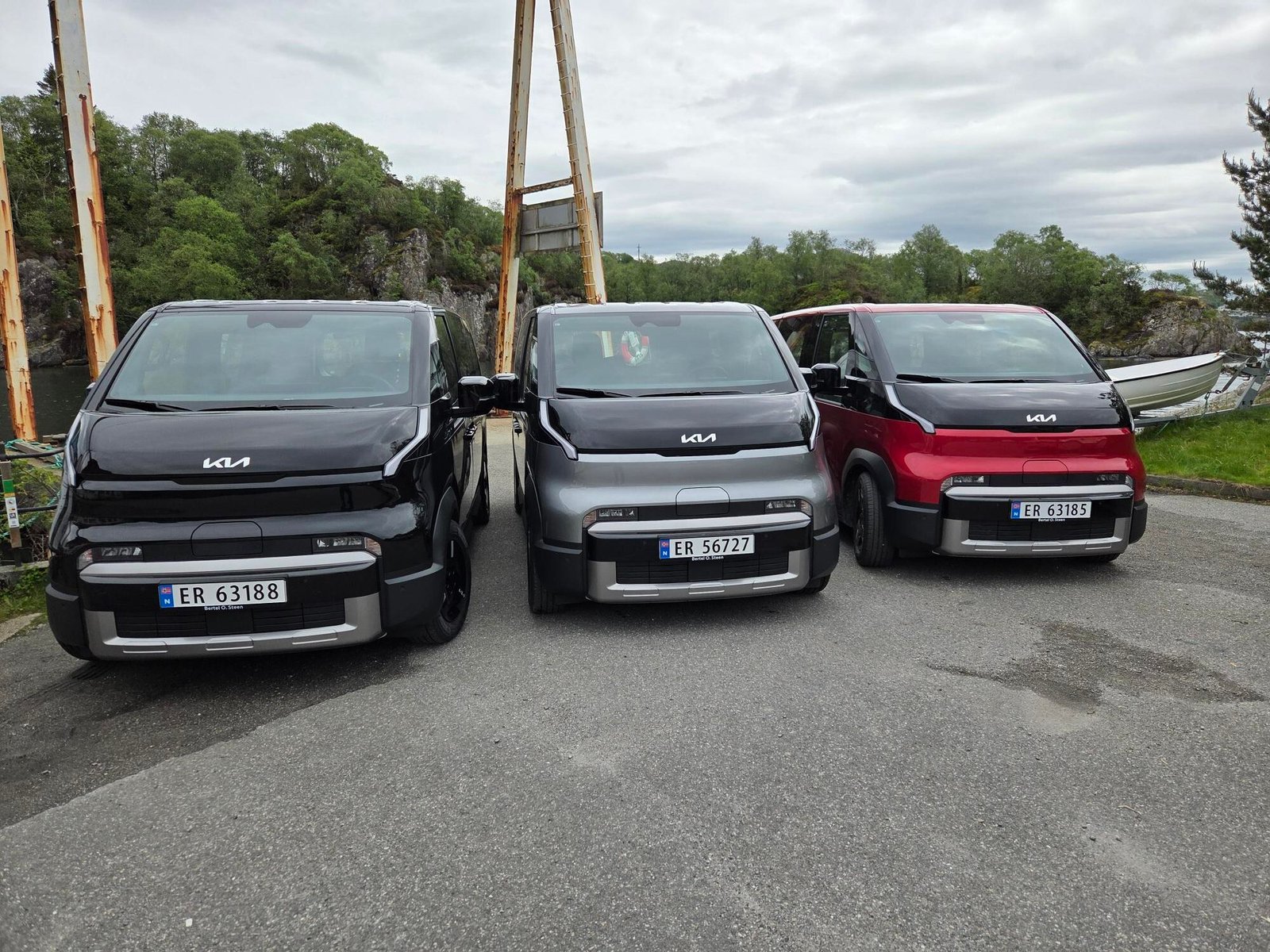 Three Kia electric vans parked in a row outdoors: black, gray, and red, with a rusty metal frame and green trees behind.